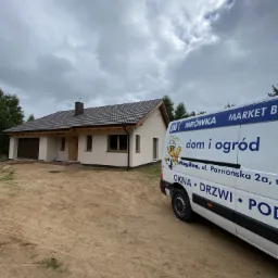 Nowo wybudowany dom z brązowym dachem i beżową elewacją, częściowo zasłonięty przez biały samochód dostawczy z logo firmy oferującej okna, drzwi i podłogi.