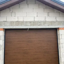 Brązowa segmentowa brama garażowa z poziomymi panelami, zamontowana w surowej ścianie z białych bloczków betonowych, widoczne ślady zaprawy.