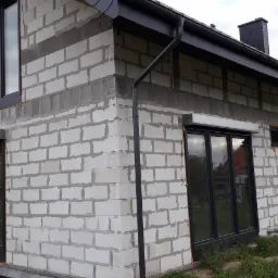 Surowy stan domu w trakcie budowy z zamontowanymi czarnymi oknami PCV i roletami zewnętrznymi, widoczne białe bloczki betonowe i elementy orynnowania.