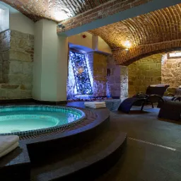 Wnętrze hotelowego SPA z jacuzzi oświetlonym od dołu, kamiennymi ścianami, ceglanym sklepieniem i leżakami z rattanu.