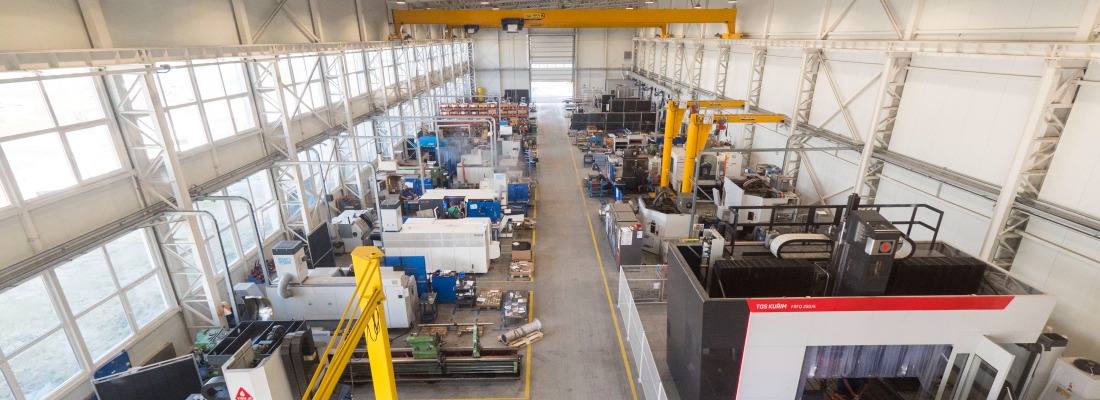 Szeroki widok hali produkcyjnej z suwnicami i różnorodnymi maszynami, w tym obrabiarkami CNC, ustawionymi wzdłuż korytarza z żółtymi liniami, oświetlonej naturalnym światłem przez okna w górnej...