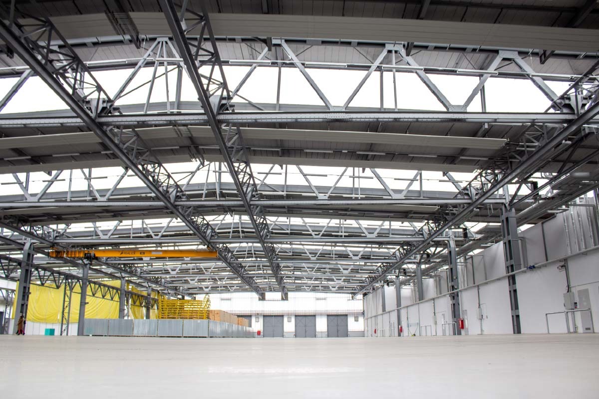 Wnętrze hali przemysłowej z widoczną konstrukcją stalową dachu, oświetleniem i suwnicą podwieszaną, żółte osłony zabezpieczające w tle.