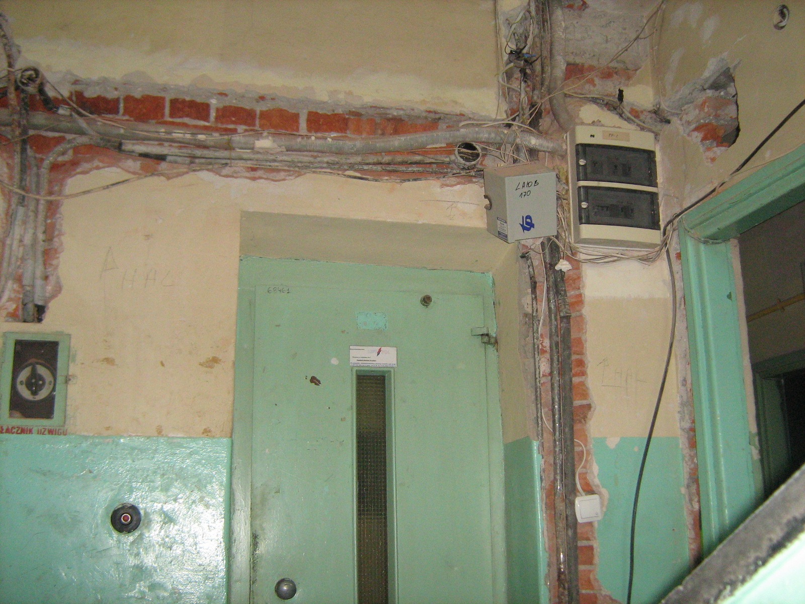 Stara klatka schodowa w budynku wielorodzinnym z widoczną tablicą rozdzielczą, przewodami elektrycznymi ułożonymi na ścianie z odsłoniętą cegłą oraz drzwiami windy z małym okienkiem.