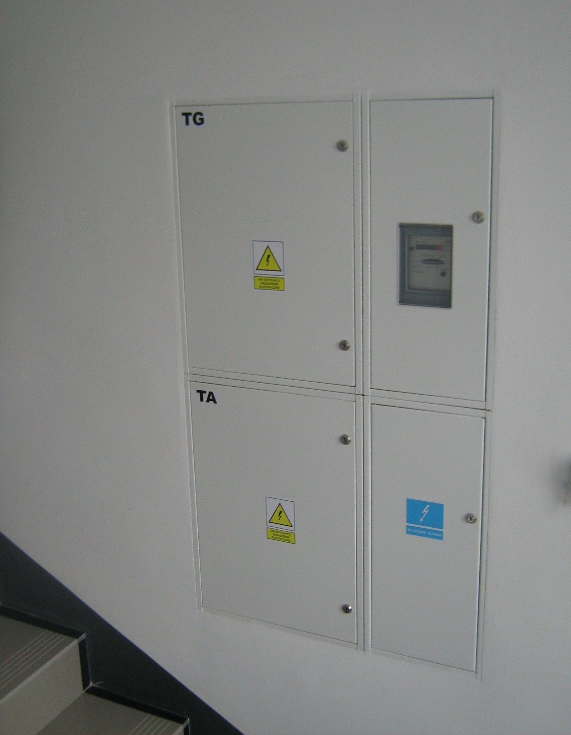 Cztery białe szafki rozdzielcze na ścianie, oznaczone TG i TA, z żółtymi naklejkami ostrzegawczymi o napięciu elektrycznym, obok schody.