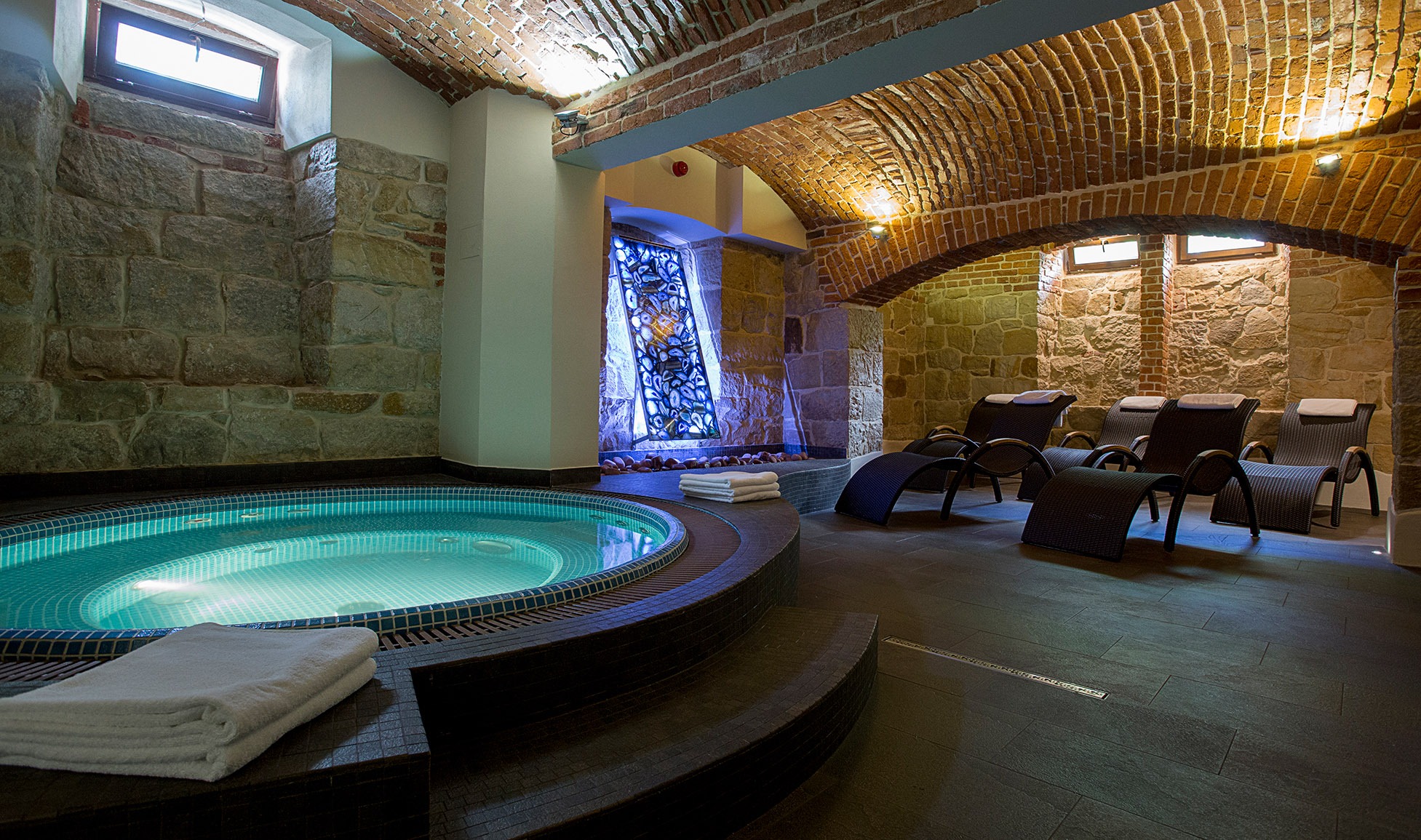 Wnętrze hotelowego SPA z jacuzzi oświetlonym od dołu, kamiennymi ścianami, ceglanym sklepieniem i leżakami z rattanu.