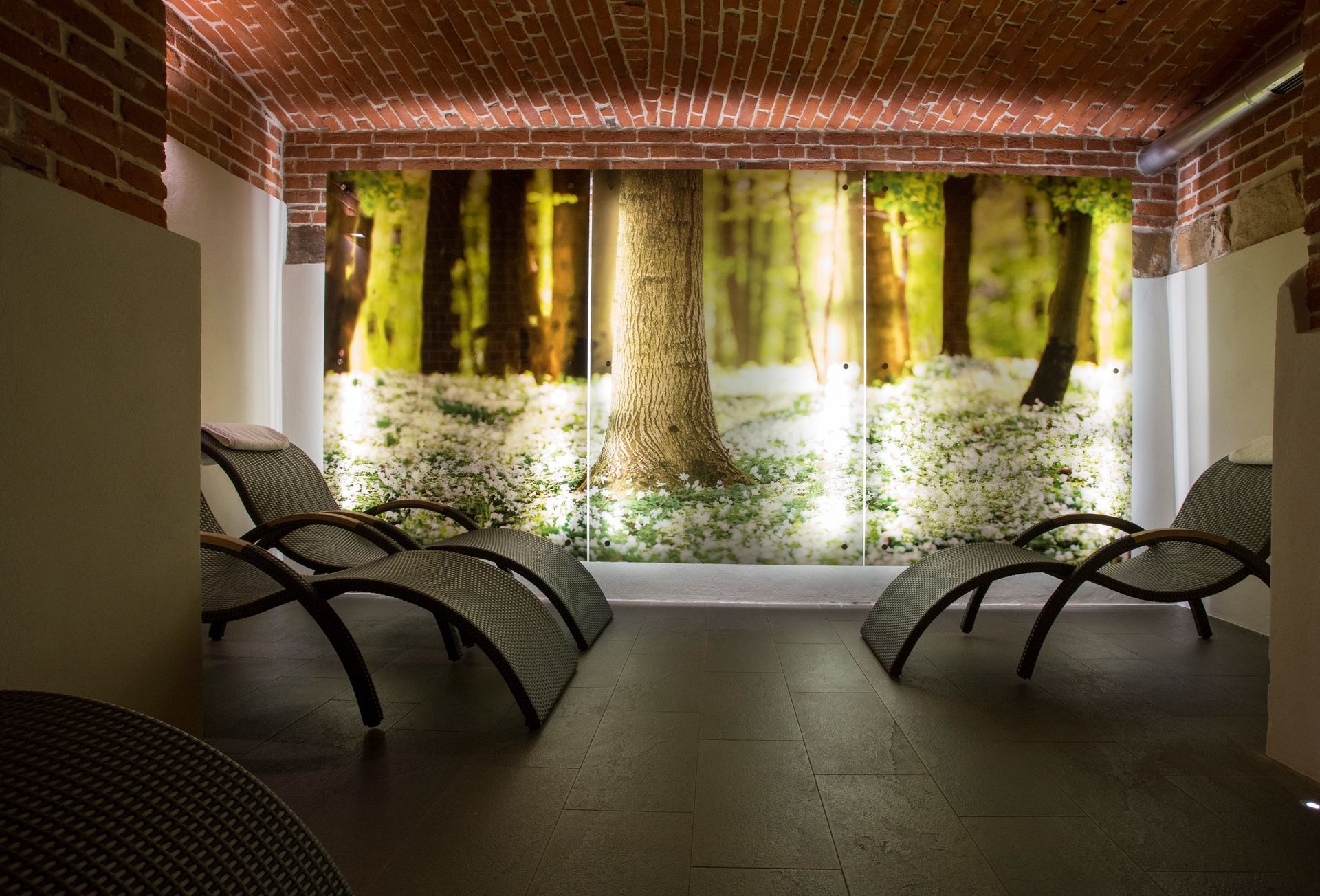 Wnętrze podziemnego SPA w hotelu: leżaki z plecionki na ciemnej podłodze, tło stanowi podświetlany obraz lasu kwitnącego na biało, sufit z czerwonej cegły.