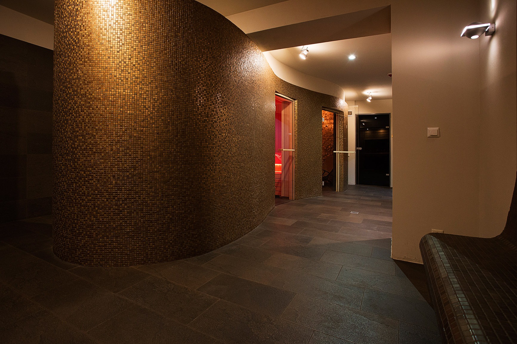 Wnętrze hotelowego SPA z zaokrągloną ścianą wyłożoną mozaiką w odcieniach brązu, ciemną podłogą z płytek, widoczne wejścia do saun i oświetlenie punktowe.