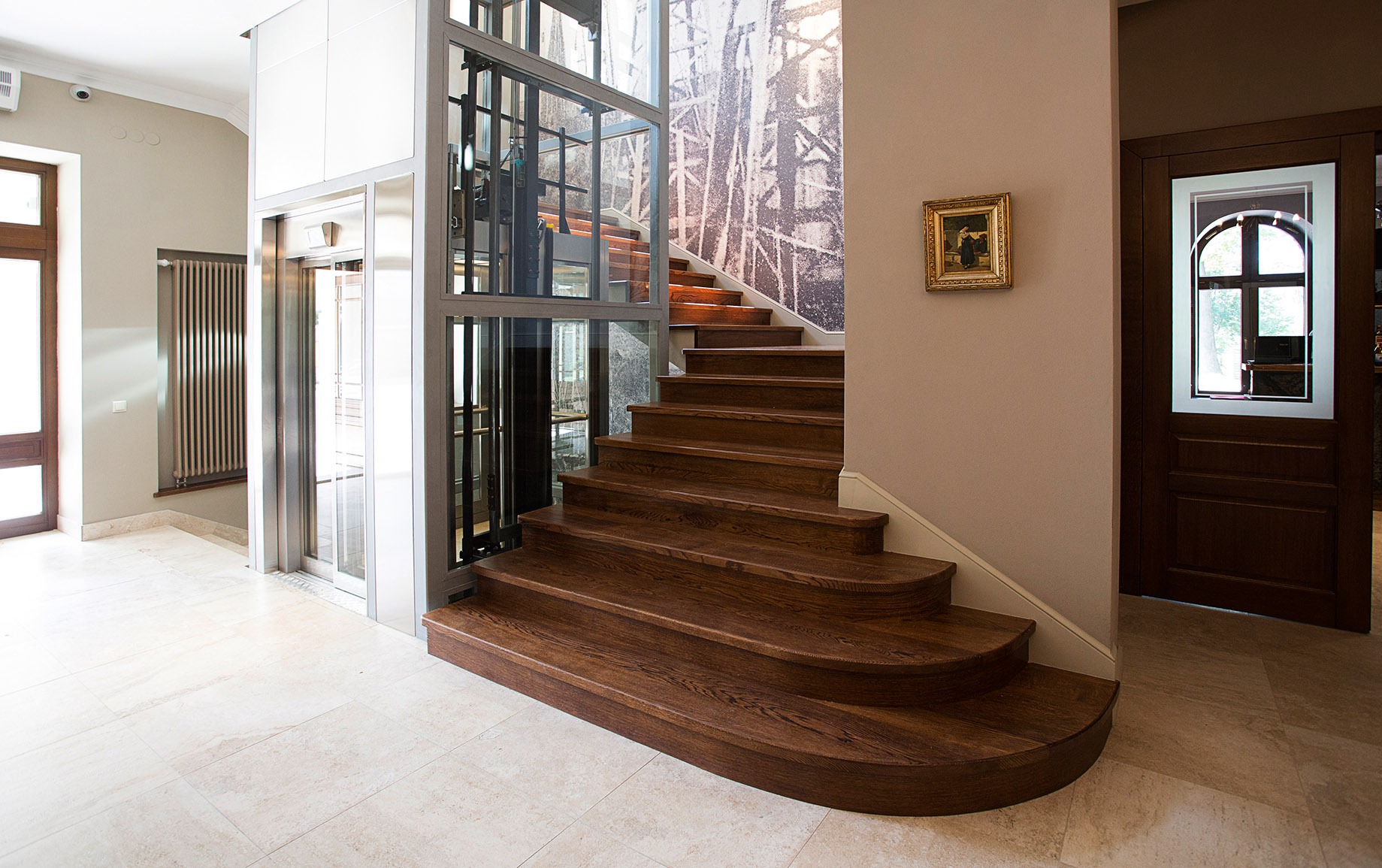 Wnętrze hotelu z drewnianymi schodami, przeszkloną windą osobową, obrazem w złotej ramie i drzwiami z okienkiem w stylu retro.