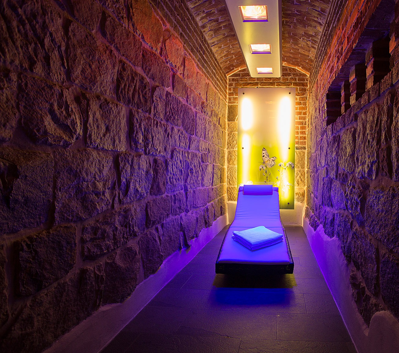 Podziemne SPA w hotelu Pałac Polanka: kamienne ściany oświetlone fioletowym światłem, leżanka z ręcznikiem, panel z motywem motyla i oświetlenie sufitowe.
