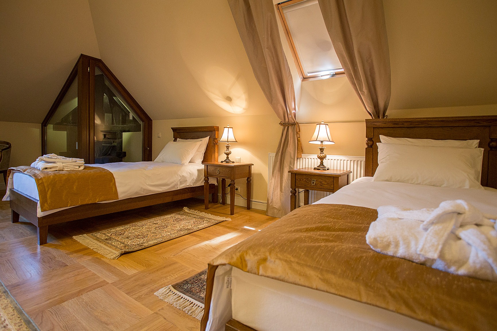 Pokój hotelowy na poddaszu z dwoma łóżkami, drewnianą podłogą w jodełkę, lampkami nocnymi i oknem dachowym z zasłonami, dodatkowo widoczny element systemu grzewczego.