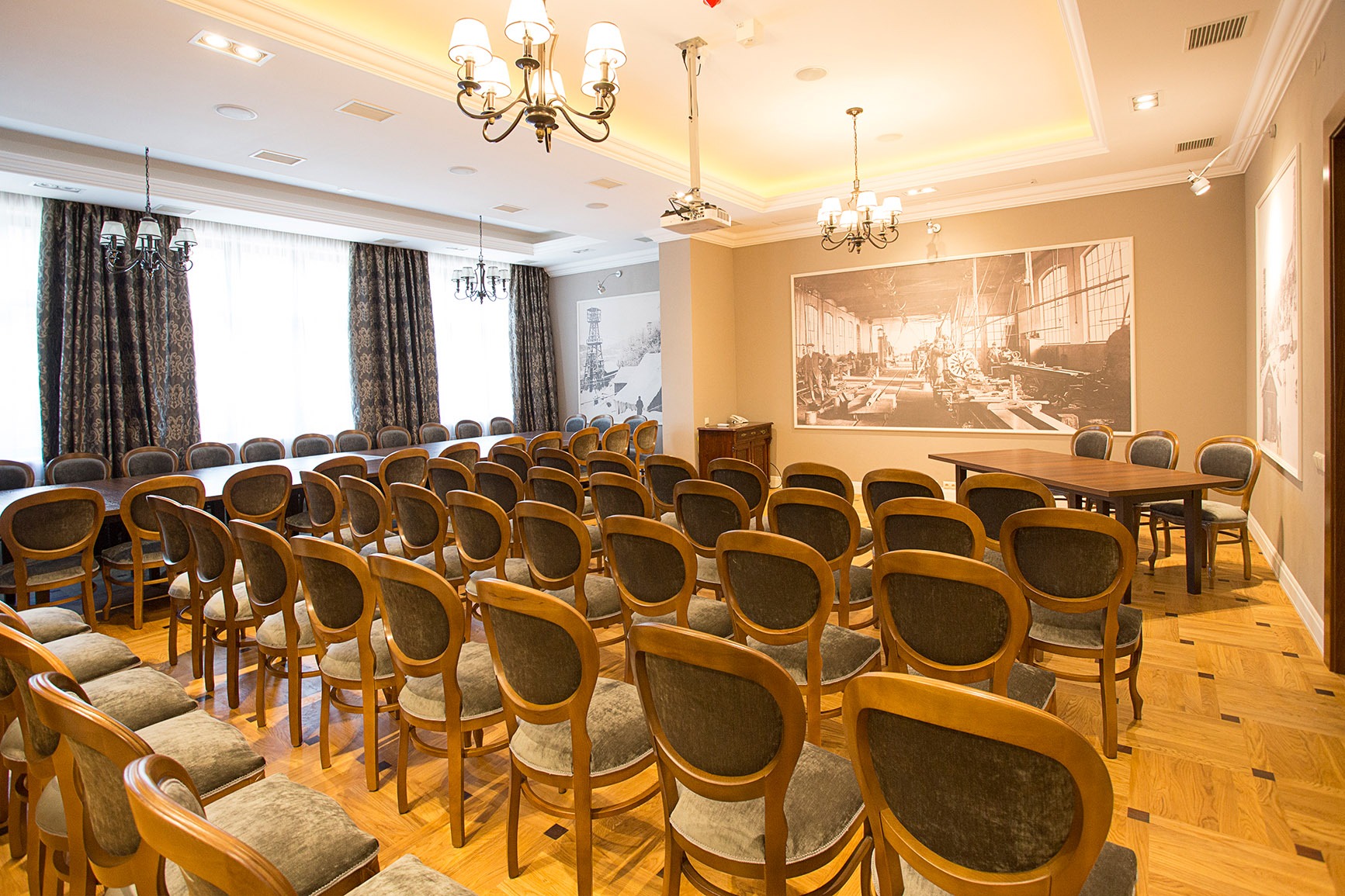 Sala konferencyjna z rzędami krzeseł i oświetleniem sufitowym, w tle projektor i duże czarno-białe zdjęcia na ścianach.