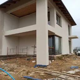 Surowy stan nowo wybudowanego domu z elewacją w trakcie prac wykończeniowych, widoczne rusztowania, teren budowy z resztkami materiałów, oraz fragment niebieskiego węża.