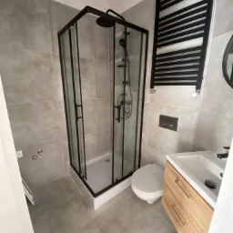 Nowoczesna łazienka z szarą, matową glazurą, kabiną prysznicową z czarnymi profilami, wiszącą toaletą, umywalką z drewnianą szafką i czarnym grzejnikiem drabinkowym.