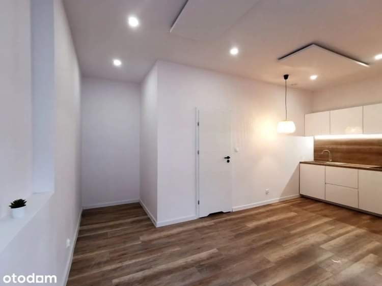 Jasne, nowocześnie urządzone wnętrze mieszkania w Czempiniu z drewnianą podłogą, białymi ścianami, minimalistyczną kuchnią i oświetleniem punktowym.
