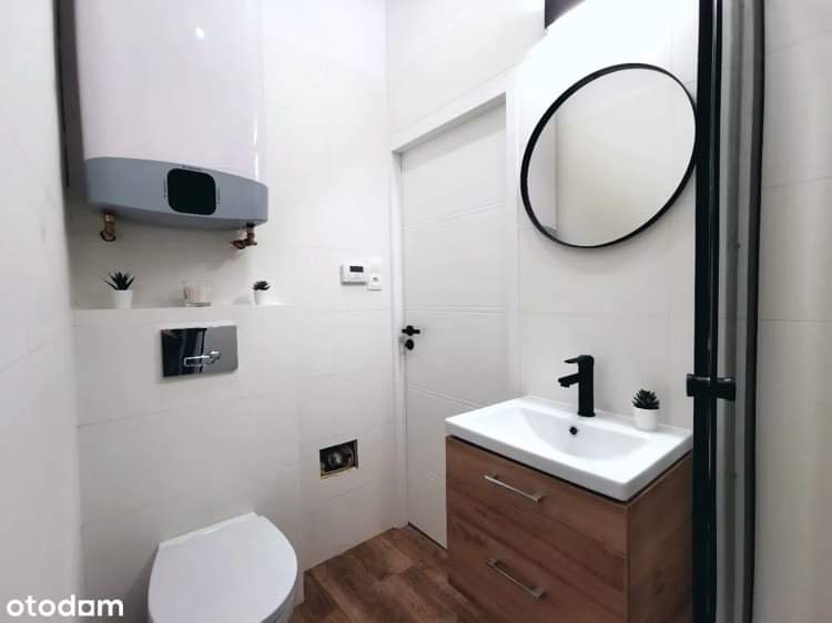 Nowoczesna, minimalistyczna łazienka z białymi płytkami, drewnopodobną podłogą, wiszącą szafką z umywalką i okrągłym lustrem w czarnej ramie; widoczny podgrzewacz wody i toaleta.