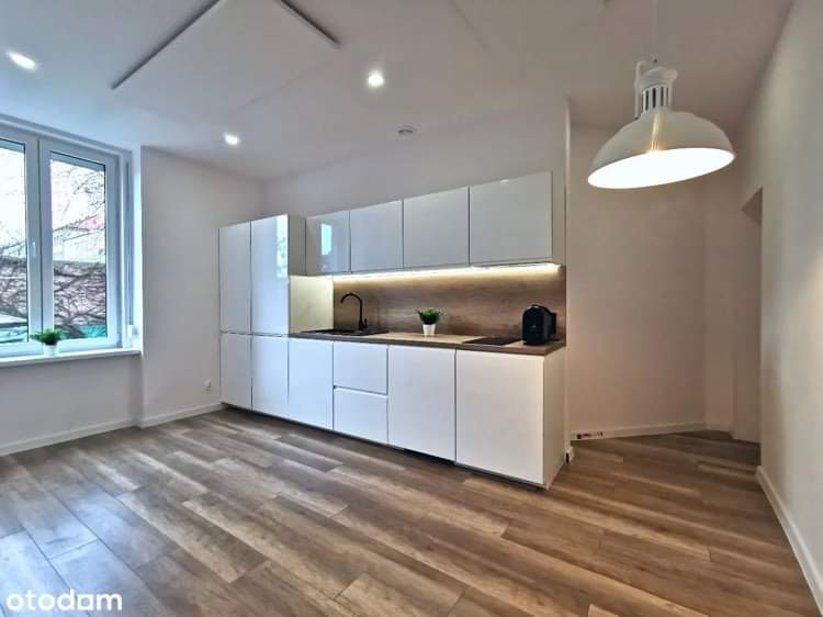 Nowoczesna, jasna kuchnia z białymi, lakierowanymi szafkami, drewnianym blatem i podświetleniem LED, widok na park przez okno, podłoga z paneli imitujących drewno ułożonych w jodełkę.