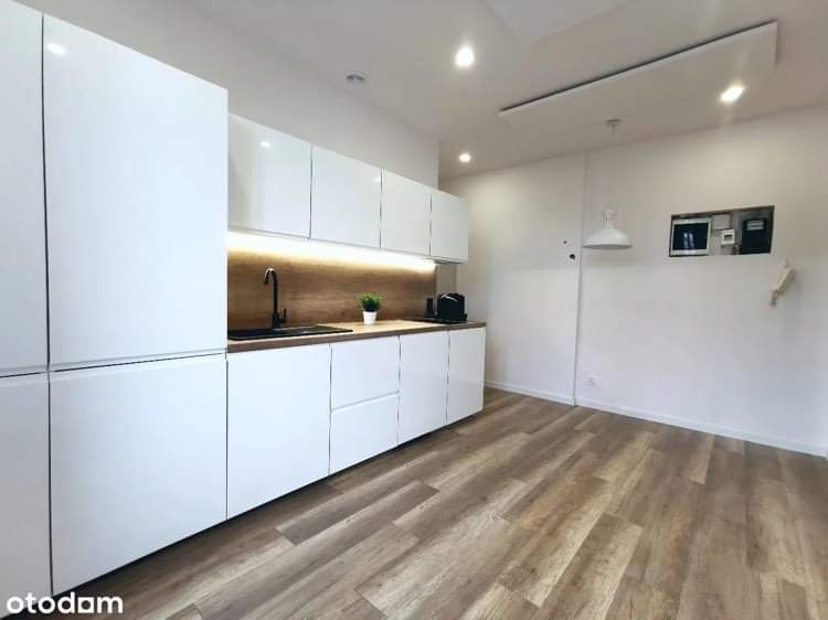 Nowoczesna kuchnia z białymi, błyszczącymi szafkami, drewnianym blatem i panelami podłogowymi ułożonymi w jodełkę, oświetlona listwą LED pod szafkami wiszącymi.