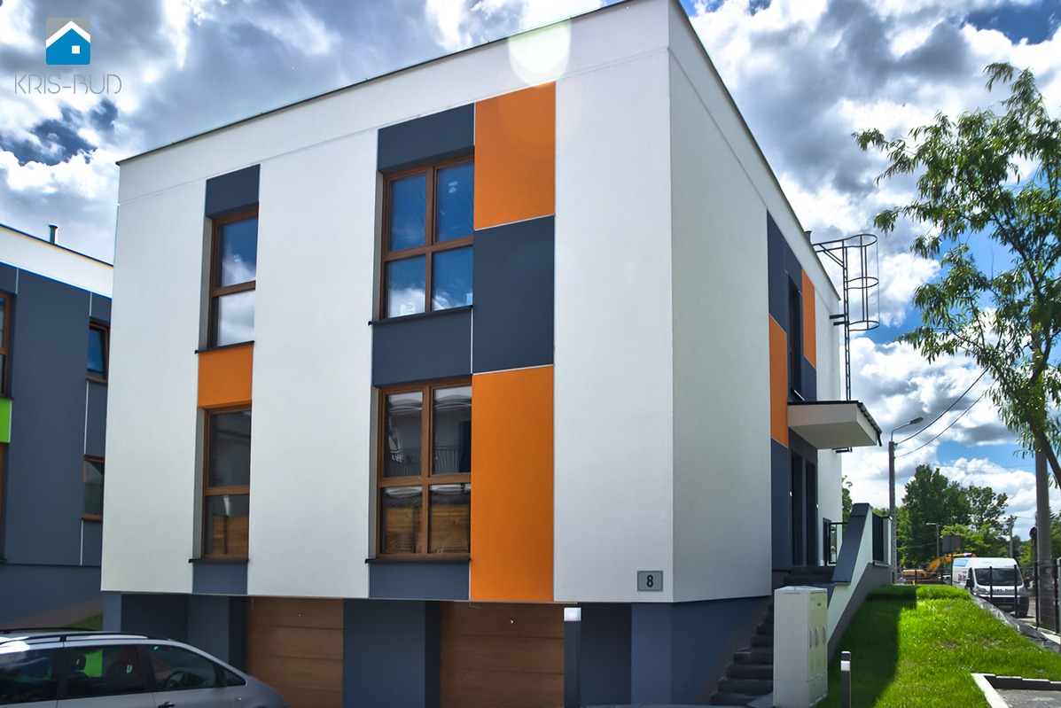 Nowoczesny, dwupiętrowy budynek mieszkalny z płaskim dachem, wykończony białym tynkiem z pomarańczowymi i grafitowymi akcentami, widoczny numer domu '8' oraz logo firmy 'Kris-Bud' w lewym górnym rogu.