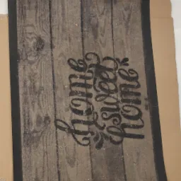 Brudny dywanik z napisem 'Home Sweet Home' przed czyszczeniem, widoczne ślady użytkowania i zabrudzenia na powierzchni.