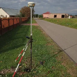 Pomiar geodezyjny w terenie zabudowanym: odbiornik GNSS na statywie, w tle droga asfaltowa, budynek w trakcie budowy i ogrodzenie z cegły.