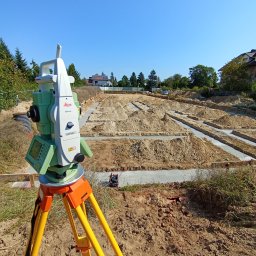 PROGEO Hubert Probola - Zielony teodolit Leica na żółtym statywie, wykopane fundamenty pod budynek w tle, błękitne niebo. Precyzyjne pomiary geodezyjne na placu budowy.