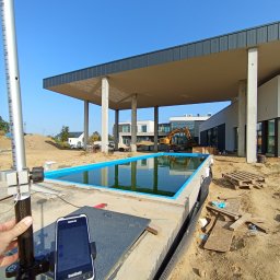 PROGEO Hubert Probola - Pomiar geodezyjny basenu w budowie. W tle budynek z zadaszeniem i koparka. Na pierwszym planie niwelator i smartfon z aplikacją pomiarową. Słoneczny dzień.