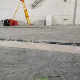 PROGEO Hubert Probola - Żółty niwelator na trójnogu w pustym pomieszczeniu z betonową podłogą. Na podłodze zielony punkt z oznaczeniem wysokości. Widoczna drabina i akcesoria.