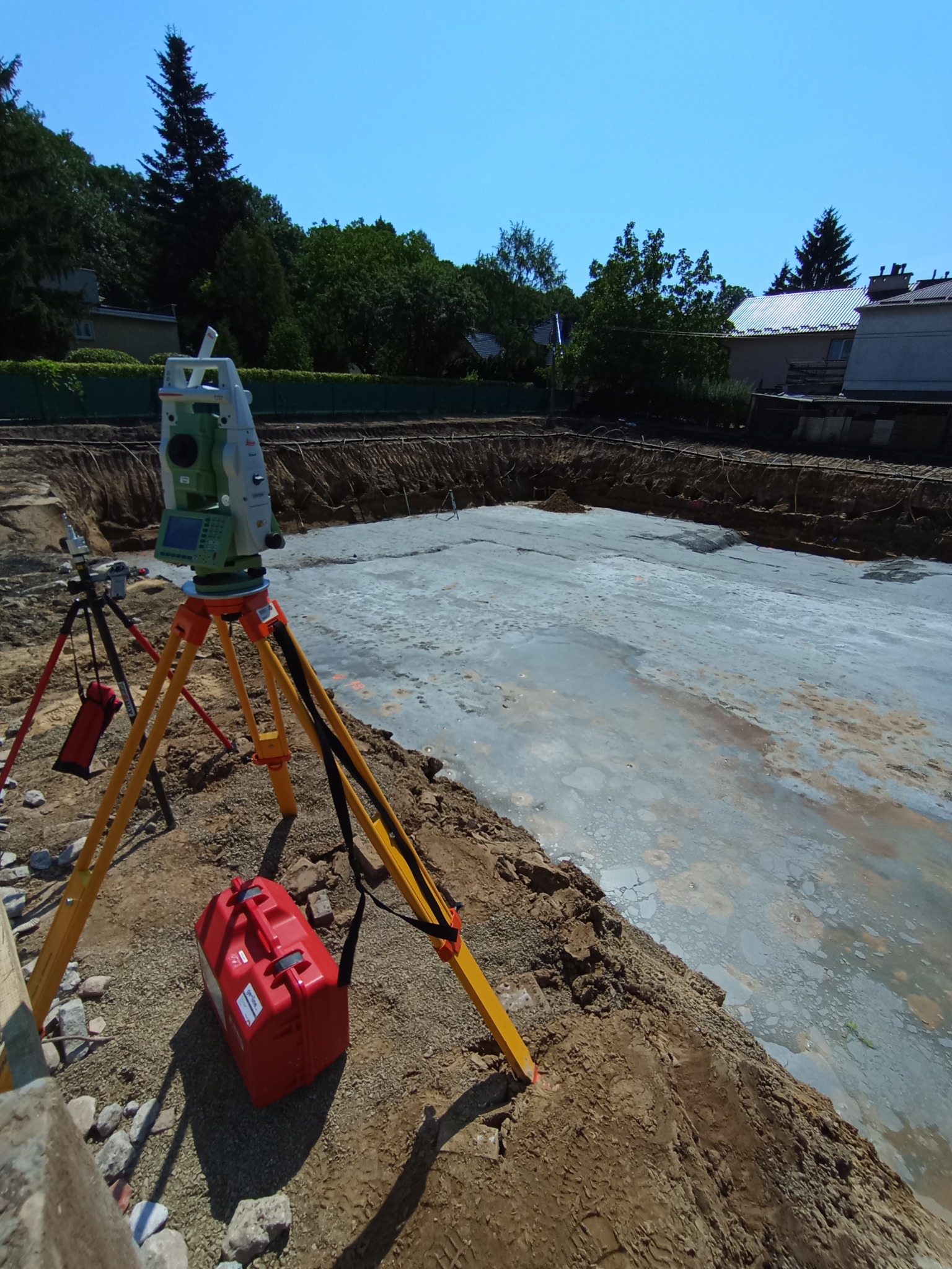 Prace pomiarowe na budowie: niwelator na statywie, wykop pod fundamenty, widok z góry. Czerwona skrzynka narzędziowa obok. Błękitne niebo w tle.