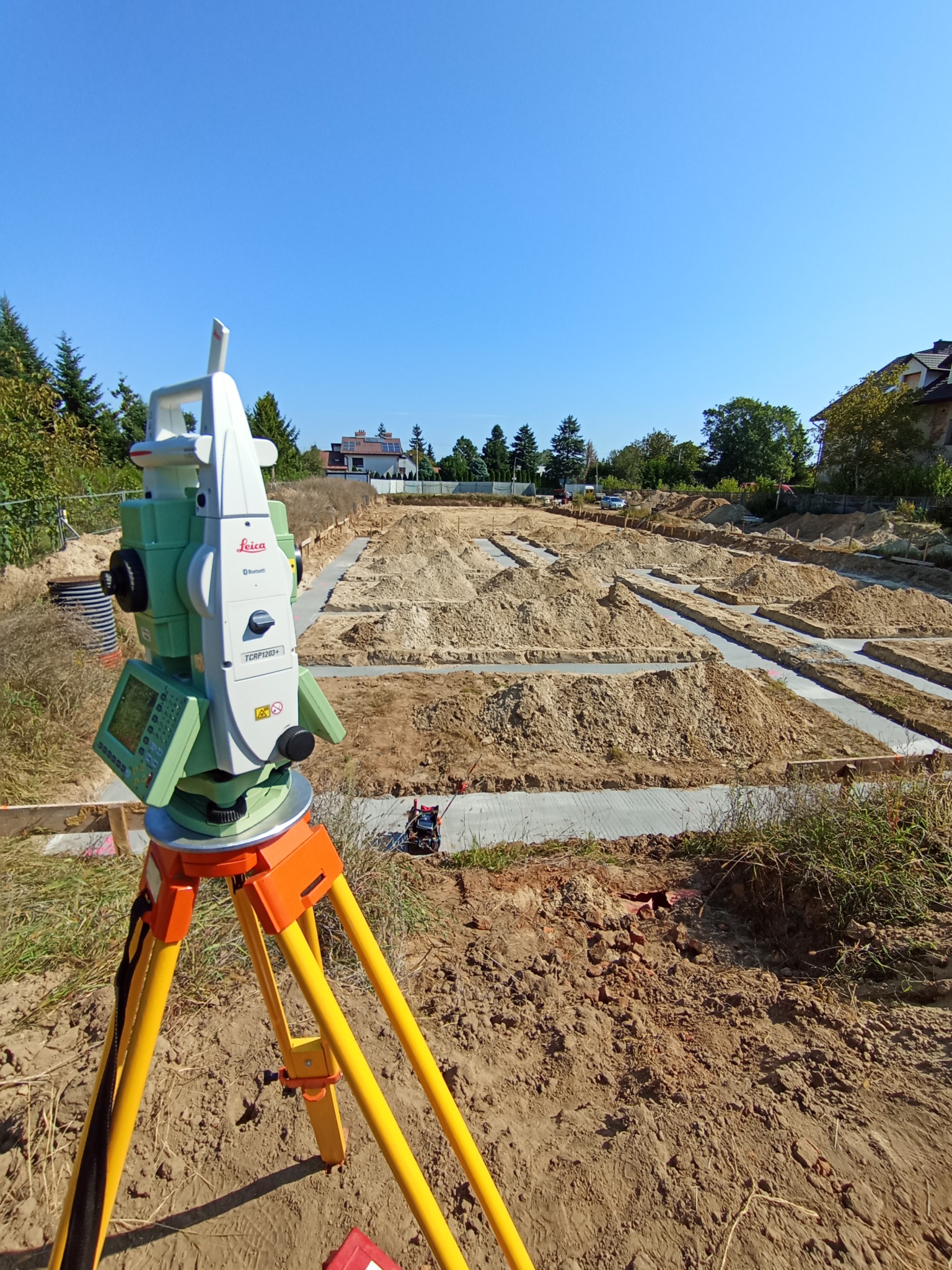 Zielony teodolit Leica na żółtym statywie, wykopane fundamenty pod budynek w tle, błękitne niebo. Precyzyjne pomiary geodezyjne na placu budowy.