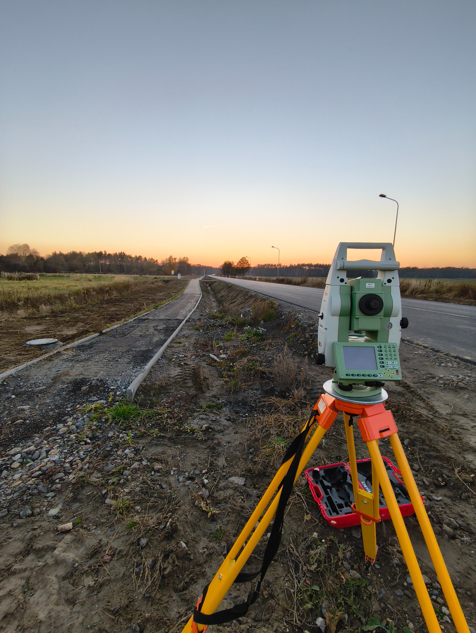 Tachimetr na statywie przy drodze w trakcie budowy, w tle zachodzące słońce. Sprzęt pomiarowy ustawiony na ziemi, obok widoczny fragment nowej ścieżki.