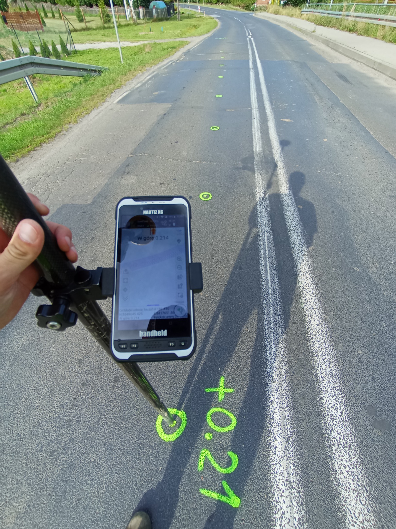 Pomiar geodezyjny drogi: smartfon na tyczce, zielone punkty pomiarowe z wartościami, cień osoby. Asfaltowa nawierzchnia z liniami. Praca geodety w terenie.