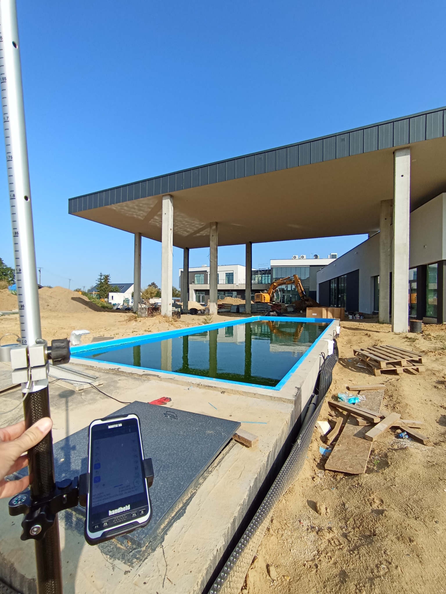 Pomiar geodezyjny basenu w budowie. W tle budynek z zadaszeniem i koparka. Na pierwszym planie niwelator i smartfon z aplikacją pomiarową. Słoneczny dzień.