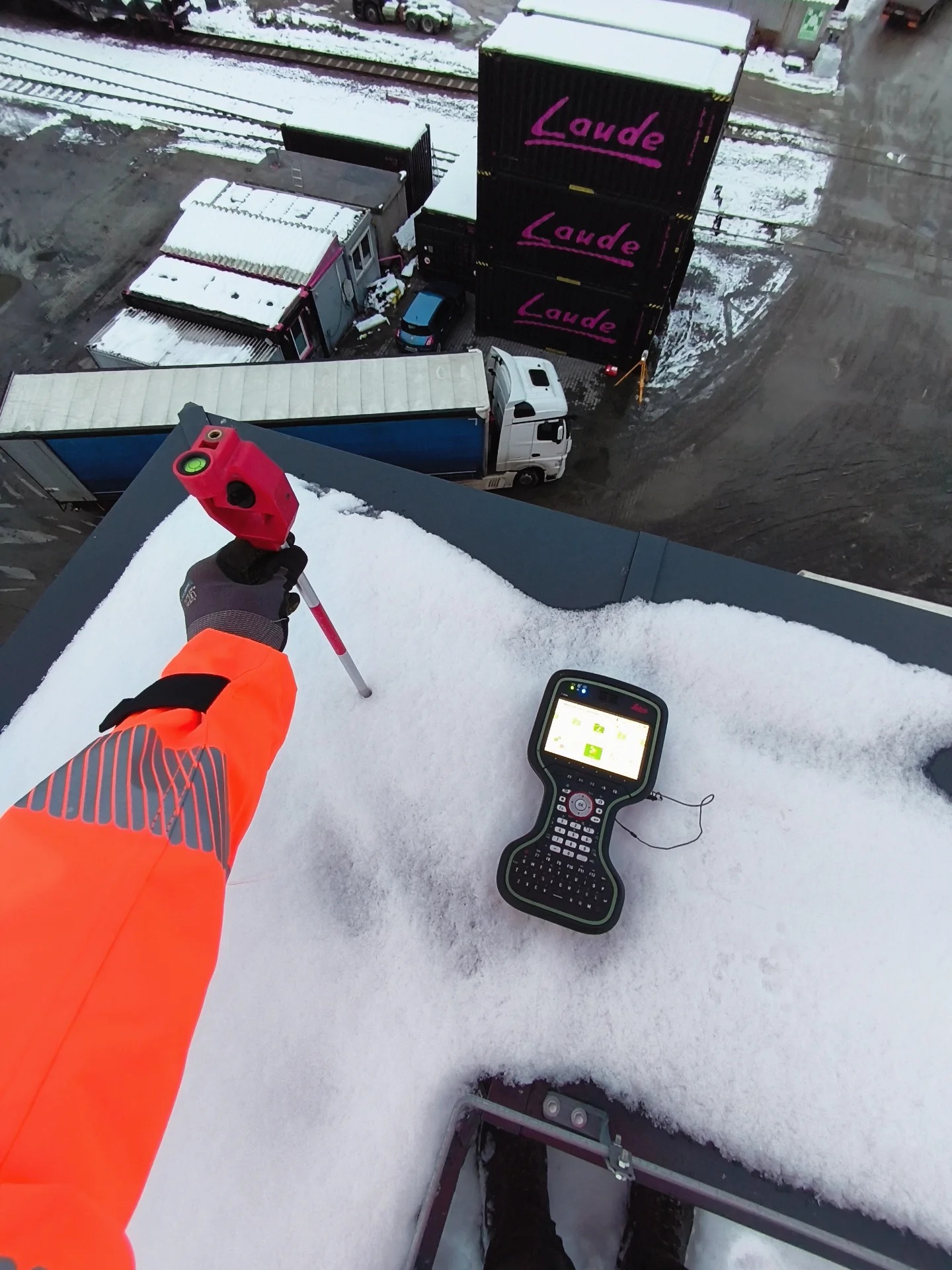 Pomiar dachu pokrytego śniegiem z użyciem niwelatora laserowego i kontrolera. Widok z góry na teren z kontenerami i pojazdami. Pomarańczowa kurtka robocza.