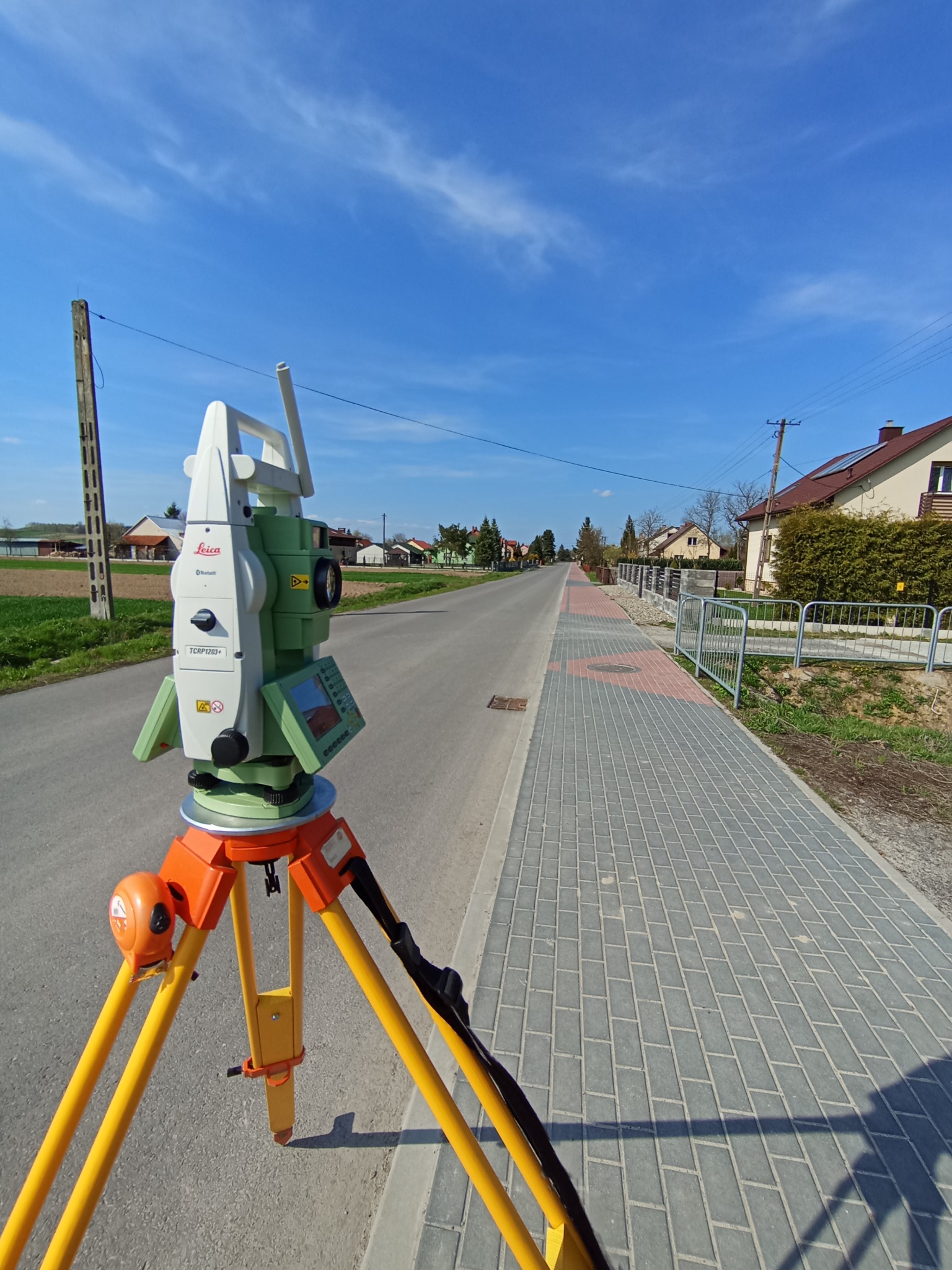 Zielono-biały teodolit Leica na żółtym statywie, ustawiony na ulicy w obszarze zabudowanym, błękitne niebo w tle. Pomiar geodezyjny w terenie.