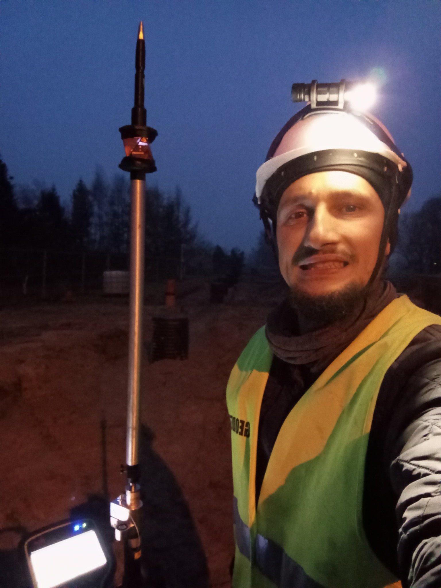 Pracownik w kamizelce odblaskowej i kasku z lampką czołową, obok tyczki geodezyjnej z pryzmatem w terenie o zmierzchu. Widoczny ekran urządzenia pomiarowego.