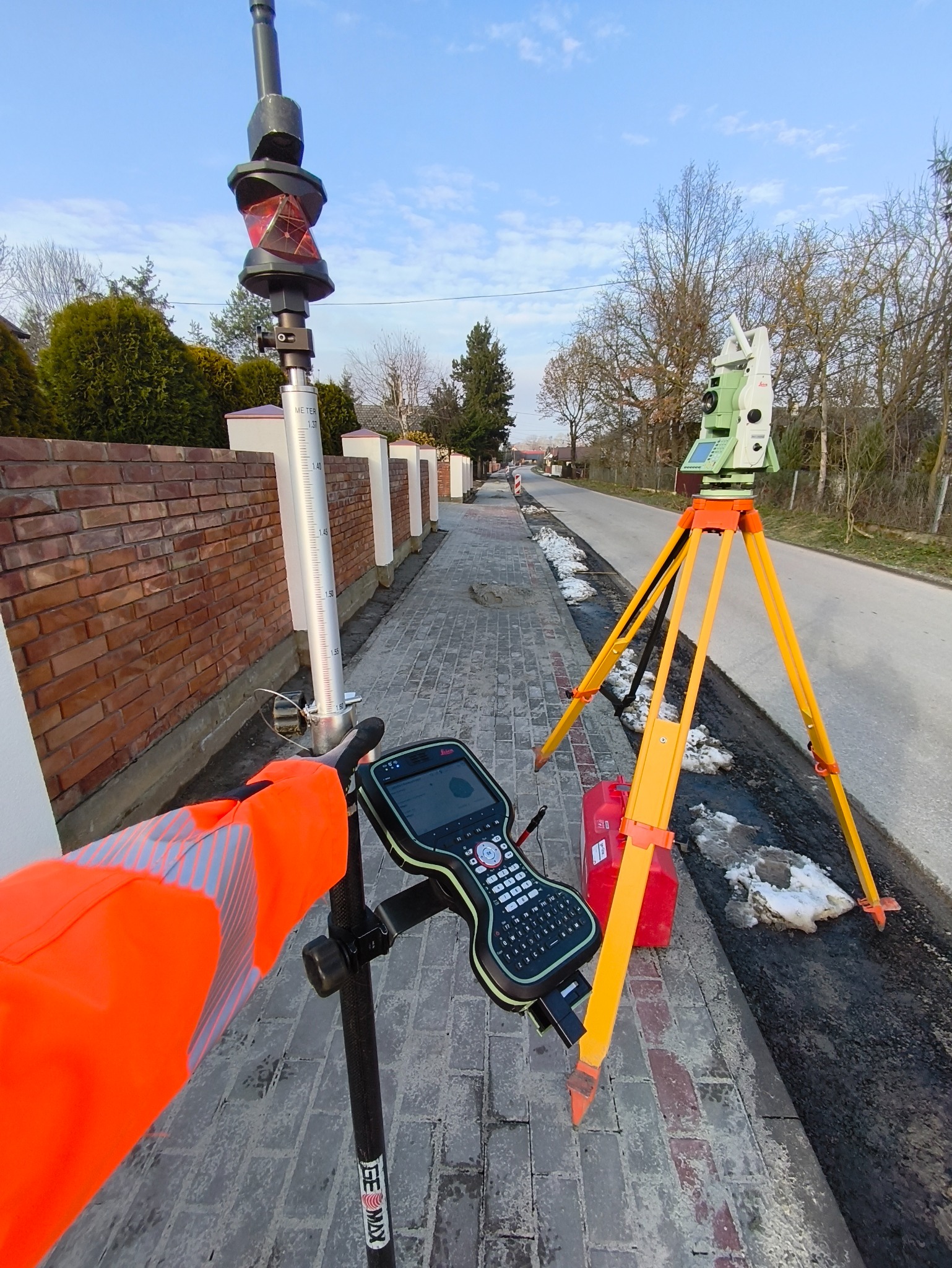 Pomiar geodezyjny: tachimetr na statywie, tyczka z pryzmatem i kontroler w pomarańczowej kamizelce na tle chodnika i ulicy.