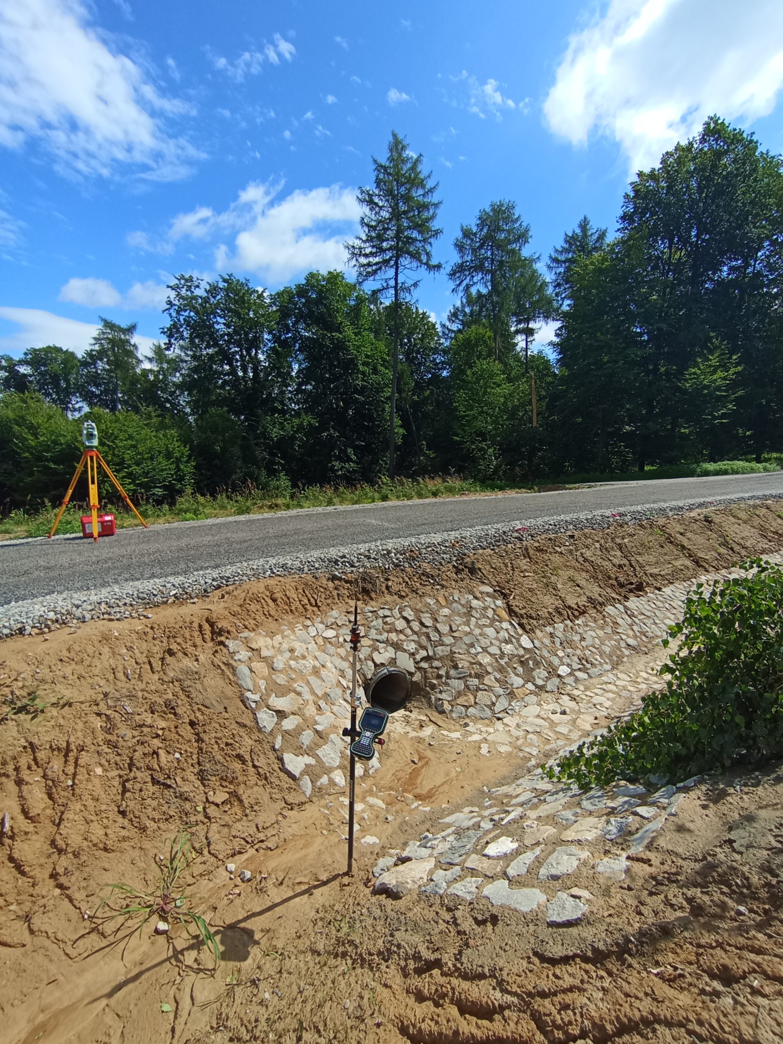 Pomiar geodezyjny nowej drogi z rowem melioracyjnym wyłożonym kamieniami. W tle drzewa i błękitne niebo z chmurami. Widoczny tachimetr i odbiornik GPS.
