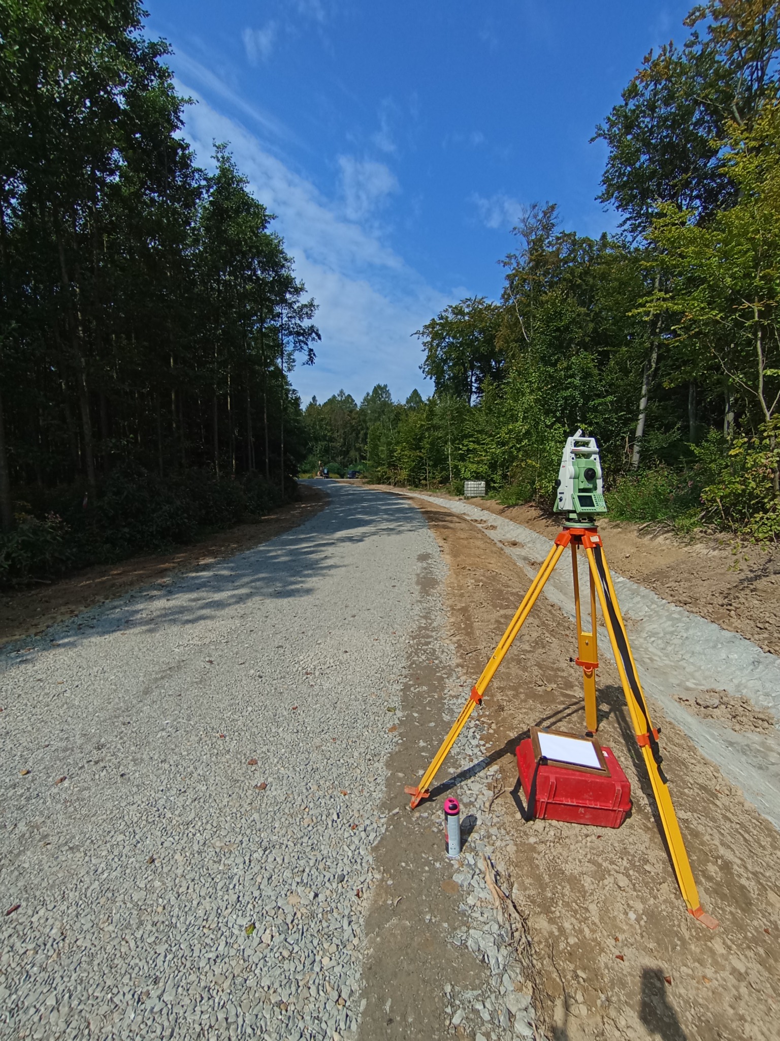 Geodezyjny teodolit na statywie, czerwona skrzynka z notatnikiem, spray obok drogi gruntowej w lesie, błękitne niebo. Prace pomiarowe w terenie.