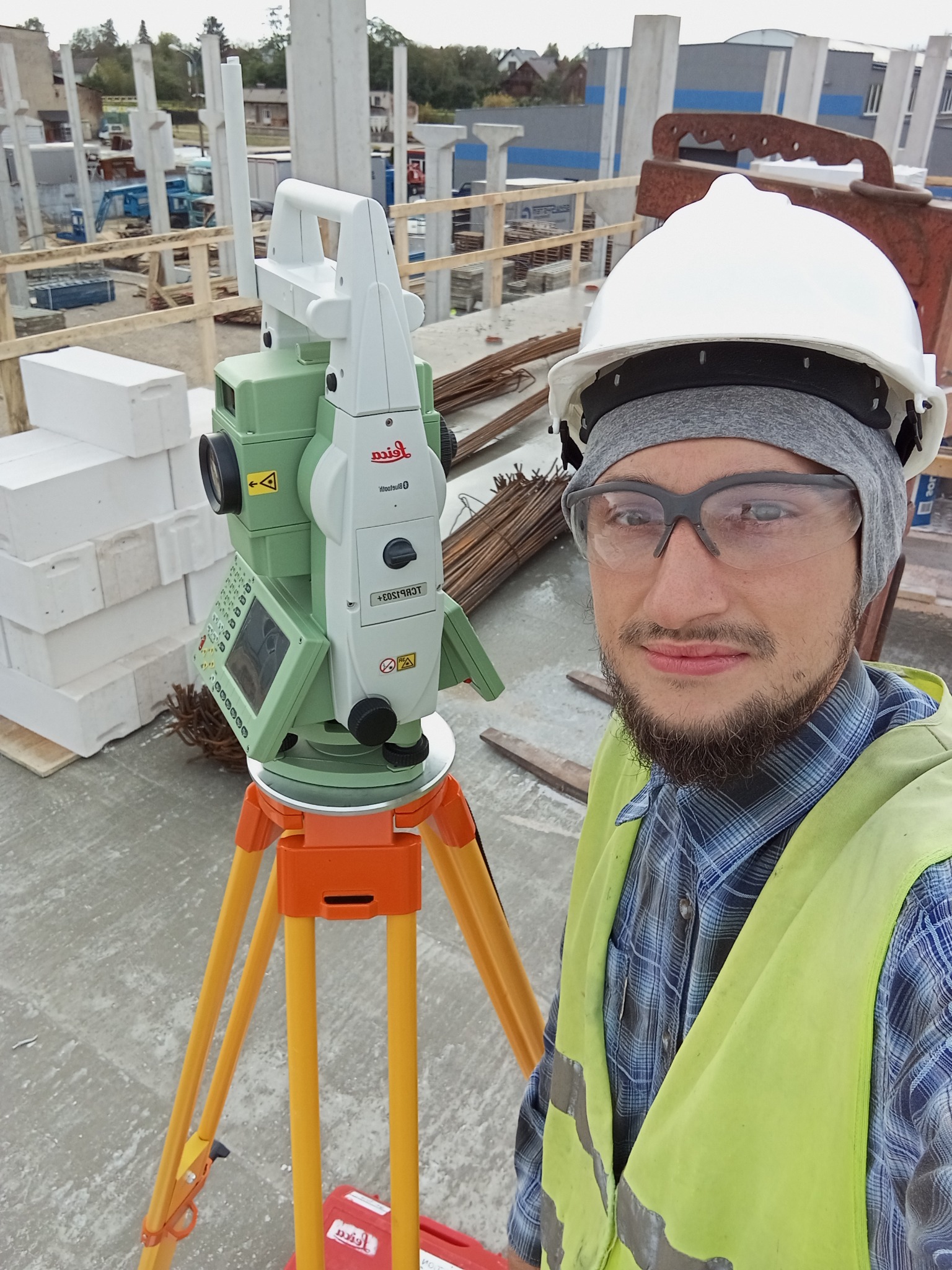 Pracownik w kasku i kamizelce obok niwelatora Leica na budowie, w tle betonowe kolumny i zbrojenia. Widok z bliska na twarz i sprzęt pomiarowy.