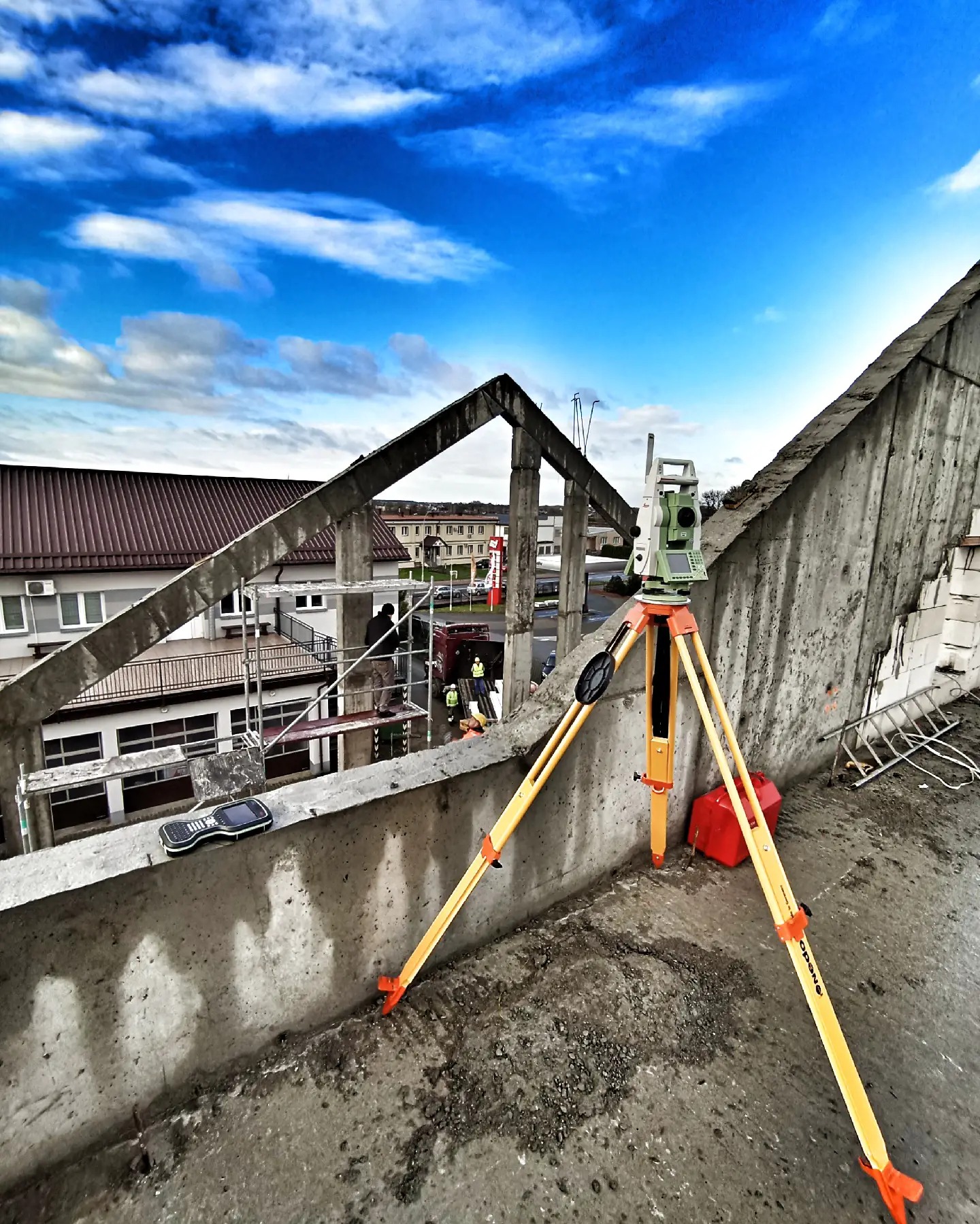 Pomiar budynku w trakcie budowy. Na pierwszym planie teodolit na tle betonowej konstrukcji i błękitnego nieba z chmurami. W tle budynek i pracownicy na rusztowaniu.