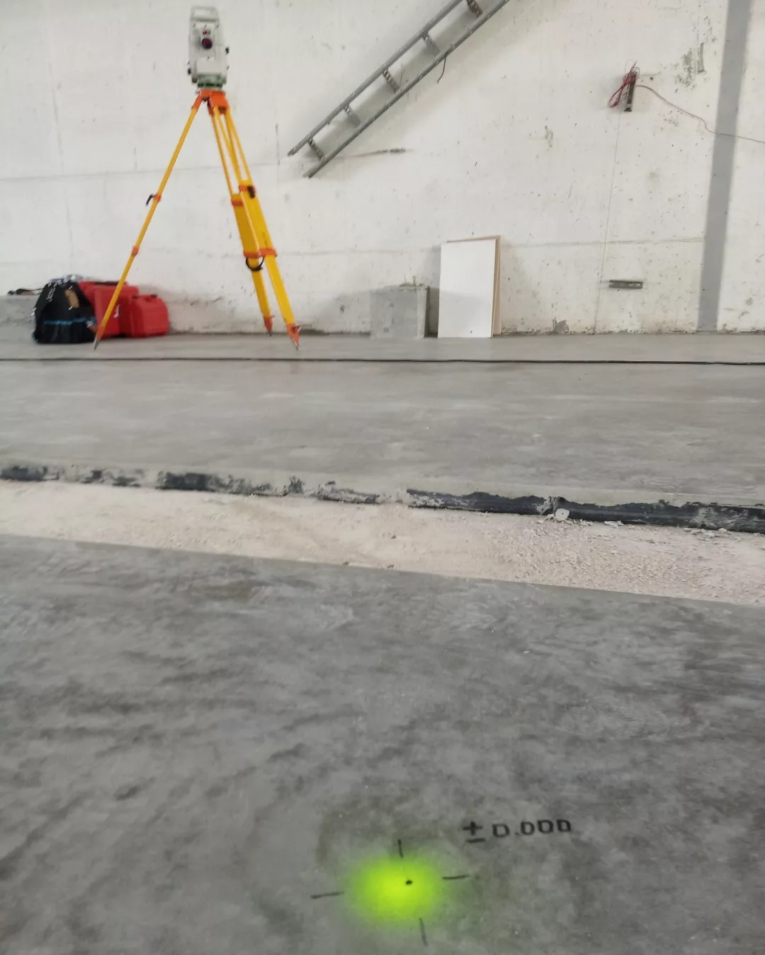 Żółty niwelator na trójnogu w pustym pomieszczeniu z betonową podłogą. Na podłodze zielony punkt z oznaczeniem wysokości. Widoczna drabina i akcesoria.