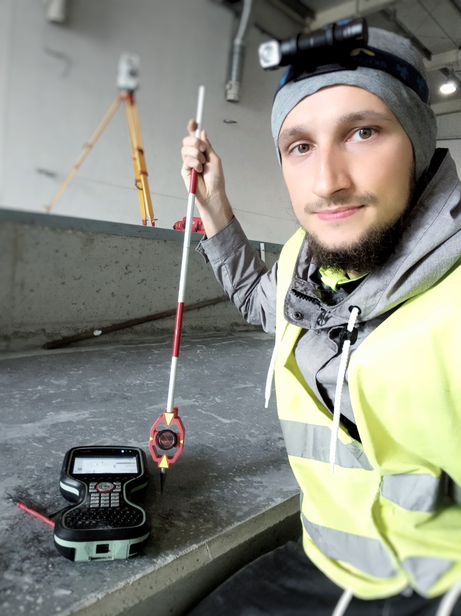 Młody mężczyzna w kamizelce odblaskowej, trzymający tyczkę geodezyjną, obok leży kontroler. W tle widoczny statyw geodezyjny i betonowa ściana.