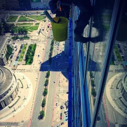 Pracownik alpinistyczny myje okna wieżowca z żółtym wiadrem, widok z góry na ruchliwą ulicę i architekturę miejską w słoneczny dzień.