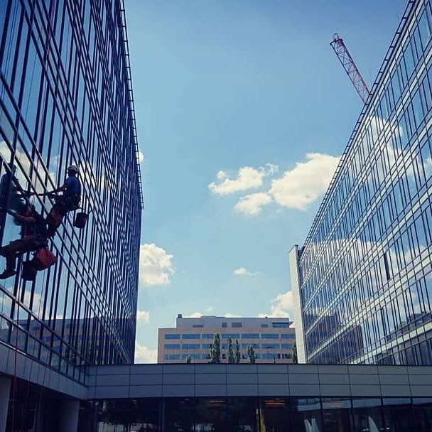 Dwóch pracowników na wysokościach, zabezpieczonych linami, czyści szklaną fasadę biurowca w słoneczny dzień. W tle widoczny inny budynek biurowy i żuraw budowlany.