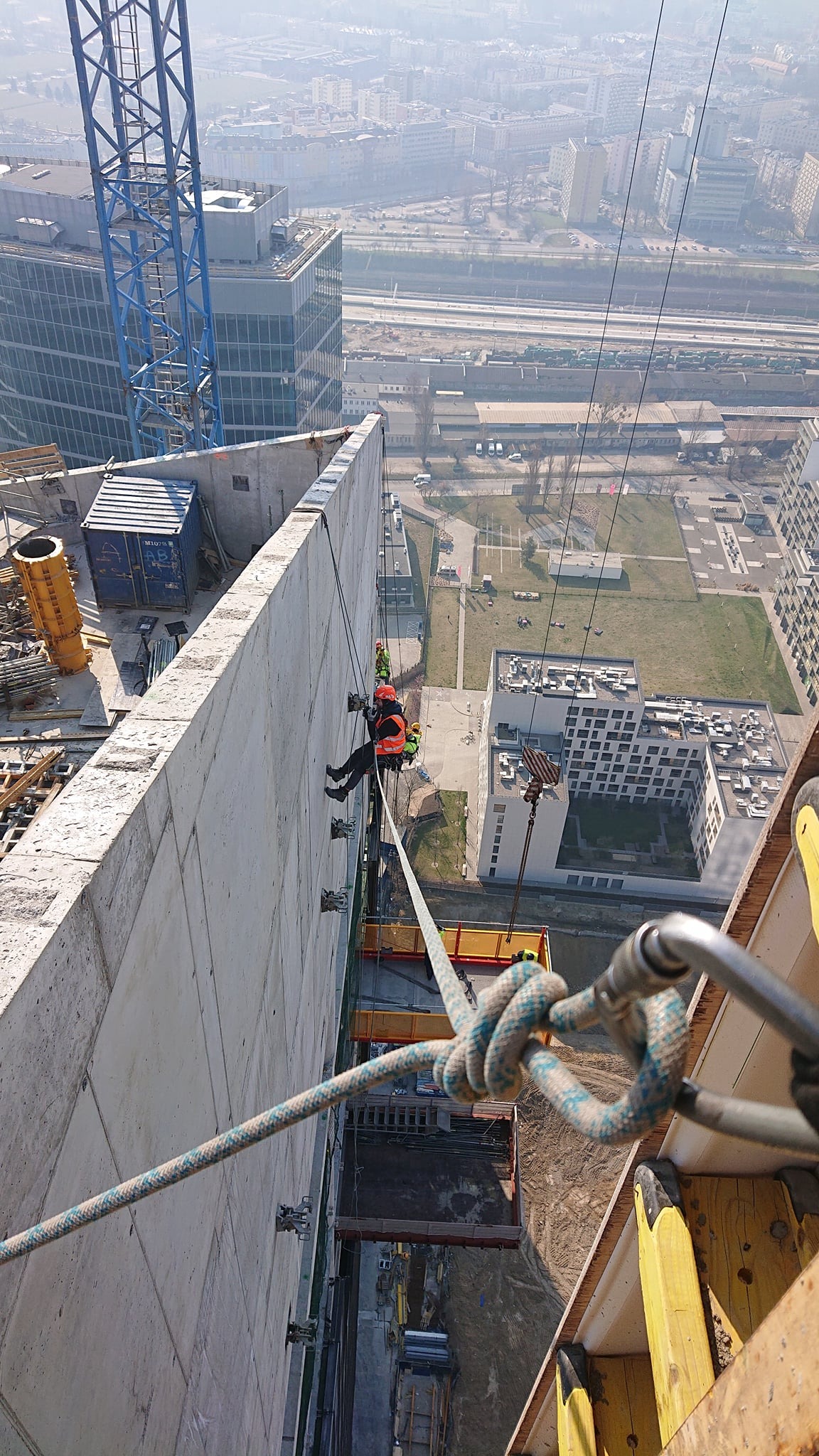 Pracownik w uprzęży, zawieszony na linie, wykonuje prace na wysokości na surowej elewacji budynku w trakcie budowy, widok z góry na miasto w tle.