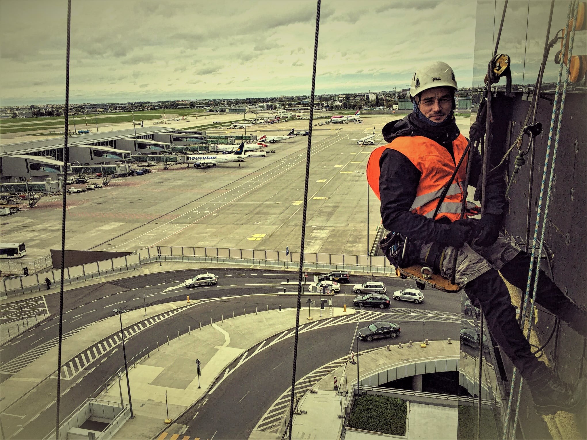 Pracownik wysokościowy w kasku i pomarańczowej kamizelce, zawieszony na linach, na tle lotniska z pasami startowymi i budynkami terminali