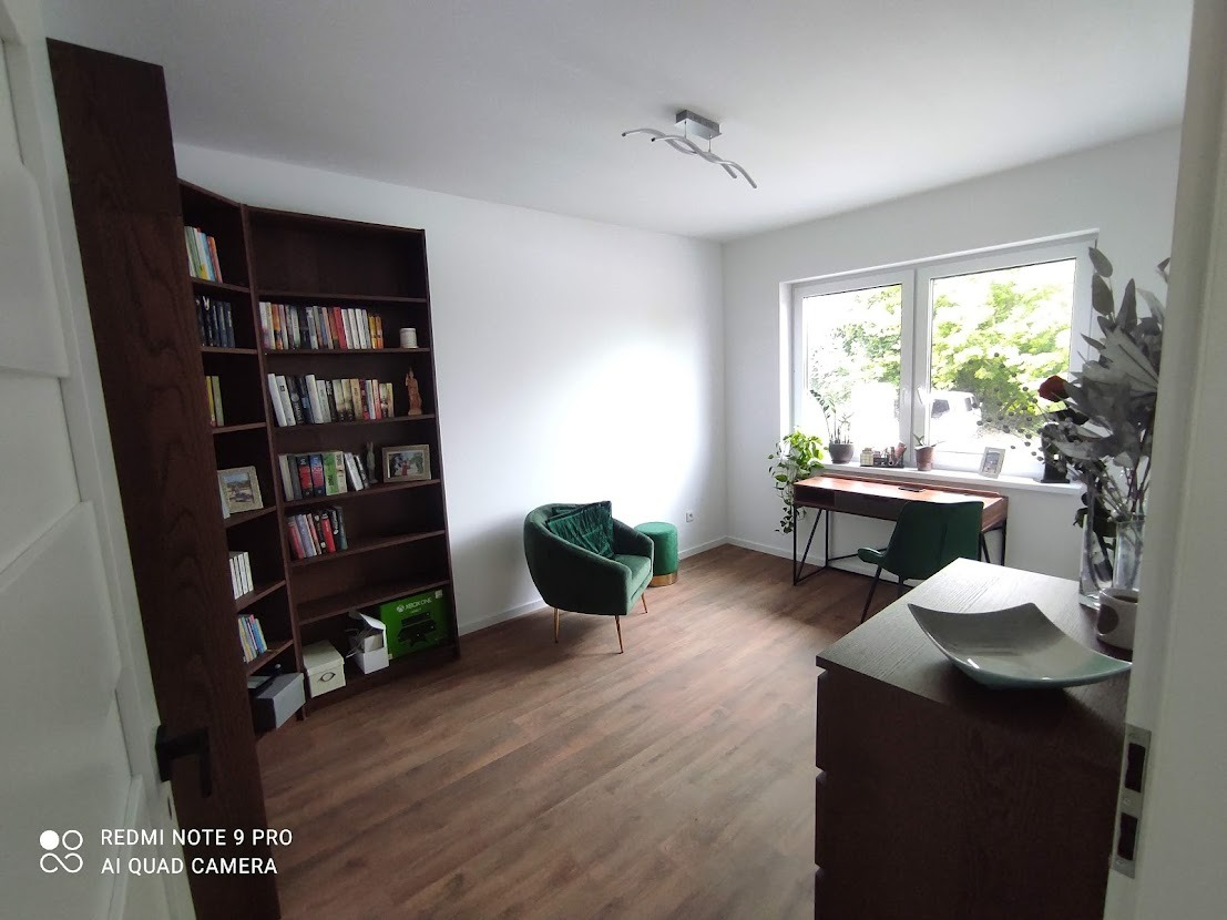 Jasne wnętrze pokoju z drewnianą podłogą, regałem z książkami, biurkiem pod oknem i zielonym fotelem z pufą, oświetlone nowoczesną lampą sufitową.