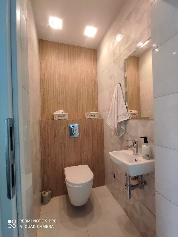 Nowoczesna toaleta z wiszącą miską WC, umywalką nablatową i geometrycznymi płytkami ściennymi, akcentem drewnopodobnych paneli i dekoracyjnymi koszykami.