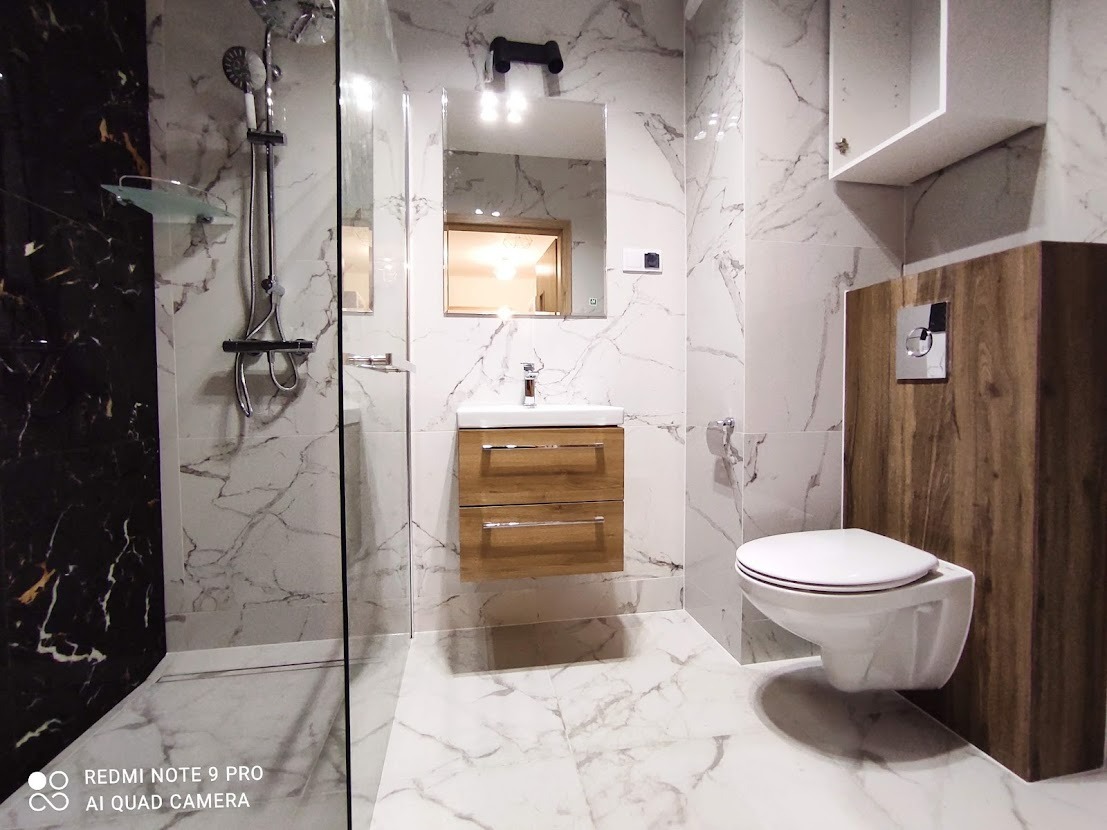 Nowoczesna łazienka z marmurowymi płytkami, kabiną prysznicową typu walk-in, podwieszaną umywalką z szafką w kolorze drewna i toaletą podwieszaną na tle drewnianej zabudowy.