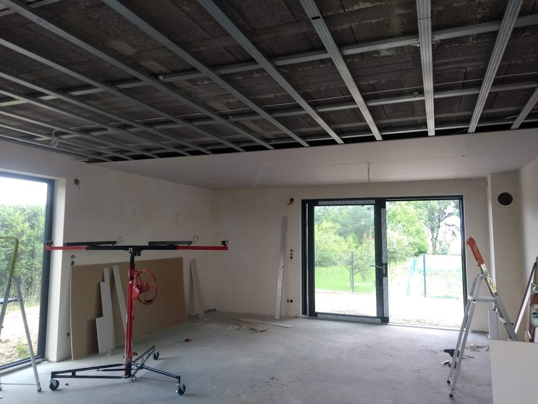 Wnętrze w trakcie montażu sufitu podwieszanego z płyt kartonowo-gipsowych, widoczna metalowa konstrukcja nośna, podnośnik do płyt z czerwoną korbą, płyty gipsowe oparte o ścianę, surowy betonowy...